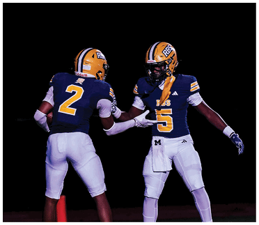 Two people in football uniforms with helmets are on a field at night, with one wearing a jersey numbered 2 and extending a hand toward the other. Description generated by AI