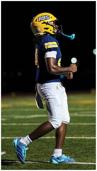A person wearing a yellow football helmet, blue jersey, and white pants is standing on a football field at night with a blue mouthguard hanging from the helmet. Description generated by AI