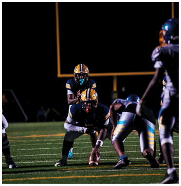 People in football uniforms are positioned on a field under stadium lights preparing for a play with a goalpost visible in the background. Description generated by AI