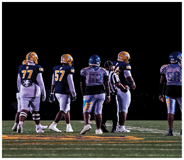 Five people in football uniforms and helmets stand on a field at night with a referee among them. Description generated by AI