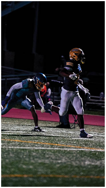 Two people in football uniforms are actively running on a field at night, with one person reaching out to grab the other. Description generated by AI