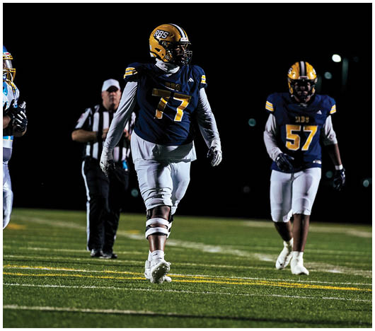 Two people in football uniforms with helmets and gloves are walking on a field at night with a referee in the background. Description generated by AI