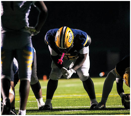 A person in a football uniform with a yellow and blue helmet is bent forward in a ready stance on a lit football field at night with other players nearby. Description generated by AI