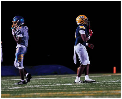 Two people in football uniforms and helmets stand facing away from each other against a dark background. Description generated by AI