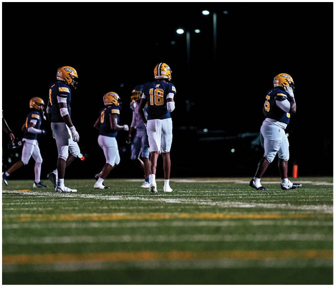 Five people wearing yellow helmets and dark jerseys stand on a football field at night under bright stadium lights. Description generated by AI