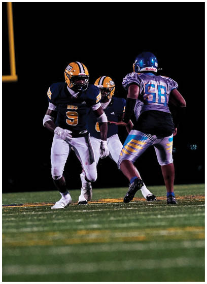 Three people in football uniforms are on a field at night, with two wearing dark jerseys and yellow helmets and one wearing a light jersey and blue helmet. Description generated by AI