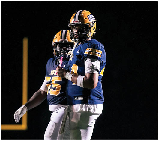 A person in a football uniform with a yellow and blue helmet is bent forward in a ready stance on a lit football field at night with other players nearby. Description generated by AI