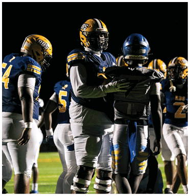 Several people wearing football uniforms and helmets are gathered on a field at night, with one person placing a hand on another's shoulder. Description generated by AI