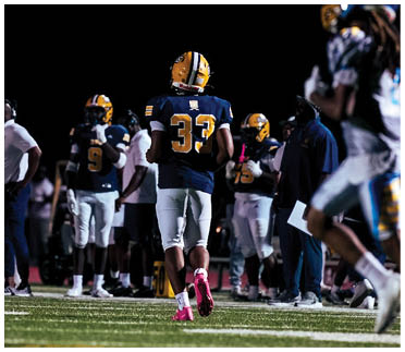 People in football uniforms are on a field at night with one person in a dark blue and yellow uniform walking away and another person in a white and blue uniform running in the foreground. Description generated by AI