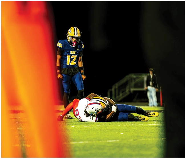 A group of people wearing football helmets and uniforms are gathered on a field under a dark night sky with a bright moon visible above. Description generated by AI