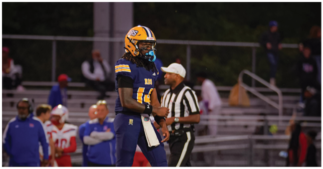 A person wearing a football uniform and helmet stands on a field with a referee and other people in the background near bleachers. Description generated by AI