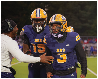 A coach wearing a headset is talking to two football players in purple and yellow uniforms on a field with trees and spectators in the background. Description generated by AI