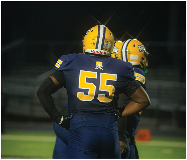 Two people wearing yellow helmets and dark blue football uniforms stand on a field with one person facing away showing the number 55 on their jersey. Description generated by AI