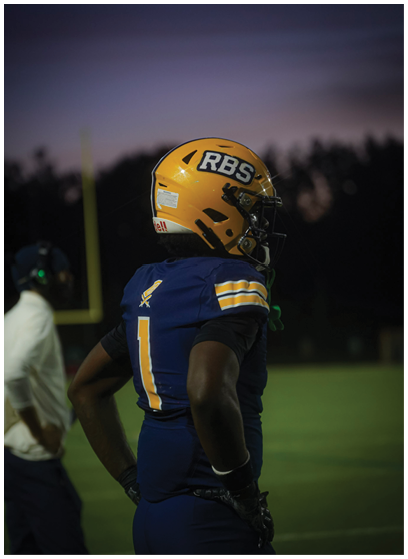 A person wearing a blue and yellow football uniform and helmet stands on a field at dusk with another person in the background near a goalpost. Description generated by AI