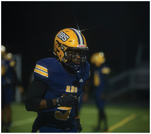 A person wearing a blue and yellow football uniform with a helmet labeled RBS is on a field at night with other players blurred in the background. Description generated by AI