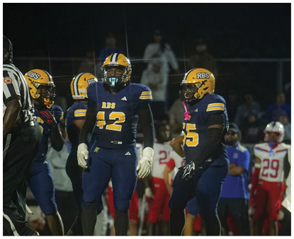 Several football players in blue and yellow uniforms stand on a field at night with other people and players in red and white uniforms visible in the background. Description generated by AI