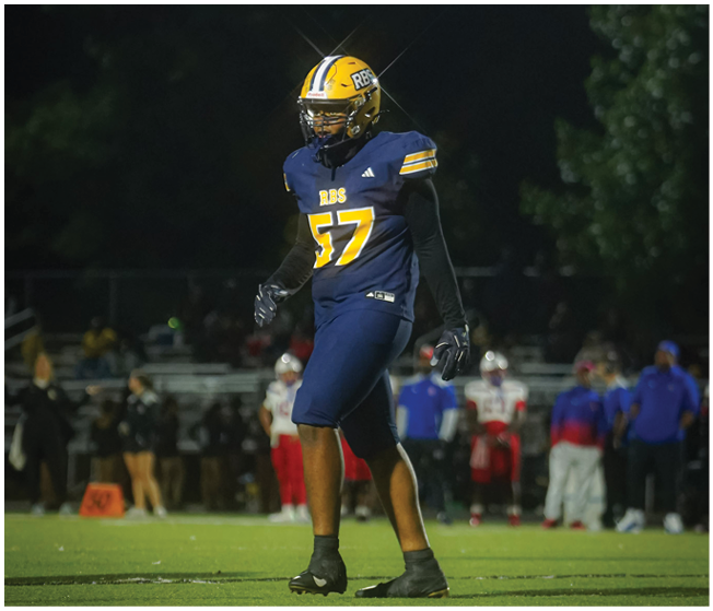 A person wearing a blue and yellow football uniform with the number 57 is standing on a field at night with trees and blurred spectators in the background. Description generated by AI