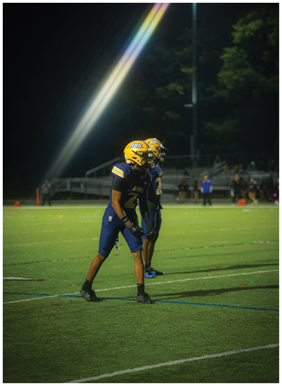 Two people wearing yellow helmets and dark blue football uniforms stand on a lit football field at night with a rainbow light streak in the sky above them. Description generated by AI