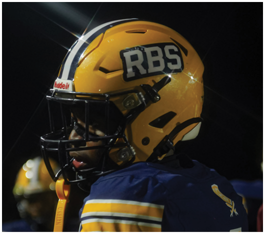 A person wearing a yellow football helmet with RBS on it and a blue jersey with yellow stripes is on a field at night. Description generated by AI