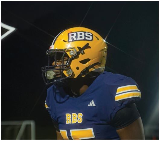 A person wearing a yellow football helmet with the letters RBS and a blue jersey with yellow and white stripes is on a field at night. Description generated by AI