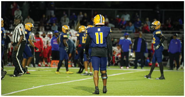 A group of people wearing football uniforms and helmets are on a field at night with spectators in the background. Description generated by AI
