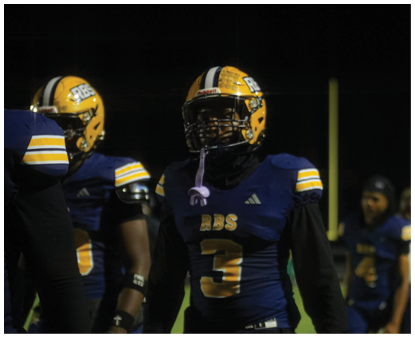 People wearing football uniforms with helmets stand on a field at night, with one person in the foreground wearing a jersey numbered 3. Description generated by AI
