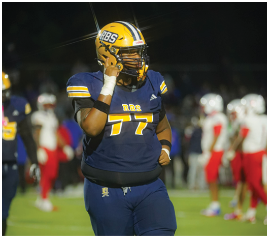 A person wearing a blue and yellow football uniform with the number 77 is standing on a field while holding a phone to their helmet. Description generated by AI