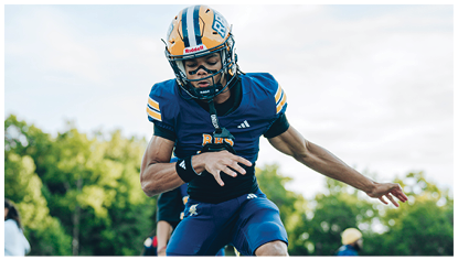 A person wearing a yellow and blue football helmet is standing outdoors with trees and a light pole in the background. Description generated by AI