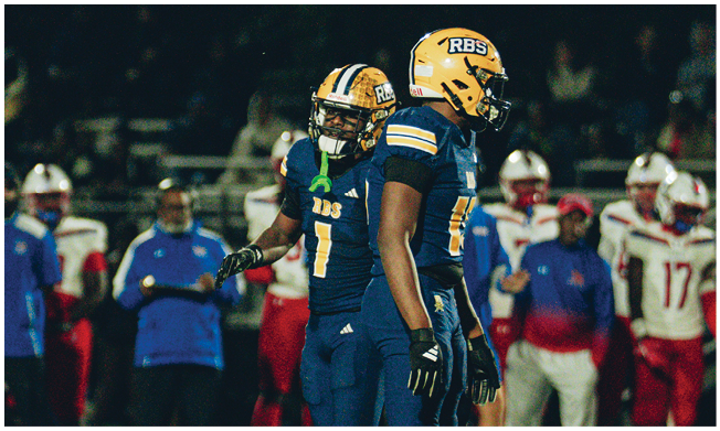 Two people in blue football uniforms stand on a field with several others in red and white uniforms in the background near an orange yard marker. Description generated by AI