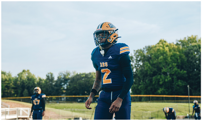 Three people wearing football helmets and uniforms stand on a field with trees and bleachers in the background. Description generated by AI