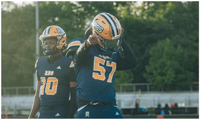 Three people wearing football helmets and uniforms stand on a field with trees and bleachers in the background. Description generated by AI