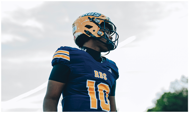 Three people wearing football helmets and uniforms stand on a field with trees and bleachers in the background. Description generated by AI