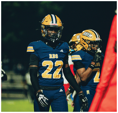 Three people wearing blue and yellow football uniforms and helmets stand on a field with trees in the background. Description generated by AI