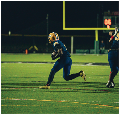 Three people wearing blue and yellow football uniforms and helmets stand on a field with trees in the background. Description generated by AI