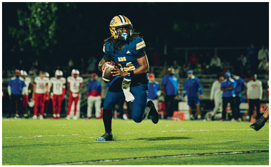 A person wearing a yellow and blue football helmet and uniform is standing outdoors under a partly cloudy sky. Description generated by AI