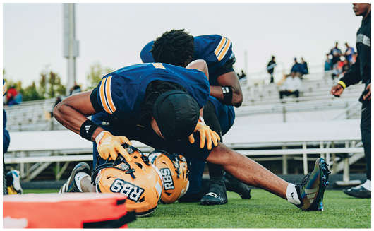 A person wearing a yellow and blue football helmet and uniform is standing outdoors under a partly cloudy sky. Description generated by AI