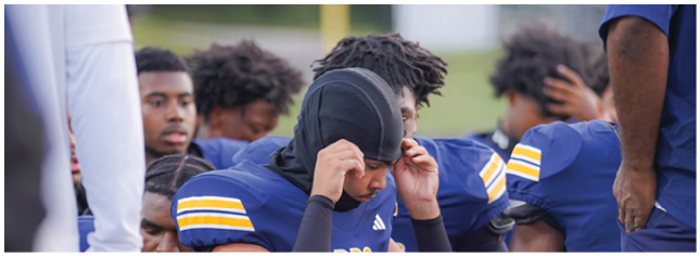 A group of people in blue sports uniforms are gathered closely, with one person adjusting a black head covering. Description generated by AI
