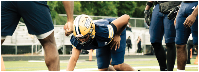 A person in football gear is crouched on the field in a ready position while other players stand nearby. Description generated by AI