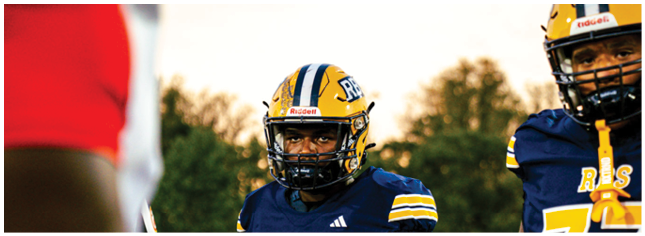Two people wearing blue and yellow football uniforms and helmets stand on a field with trees in the background. Description generated by AI