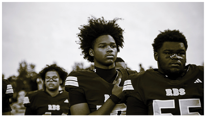Three people wearing football uniforms with face paint stand closely together outdoors, with one person placing a hand on another's shoulder. Description generated by AI