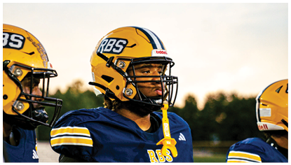 Three people wearing yellow helmets and navy blue football uniforms stand outdoors with a blurred background of trees and sky. Description generated by AI