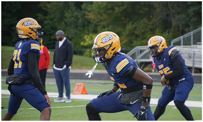 Three people wearing football helmets and uniforms stand on a field with trees and bleachers in the background. Description generated by AI