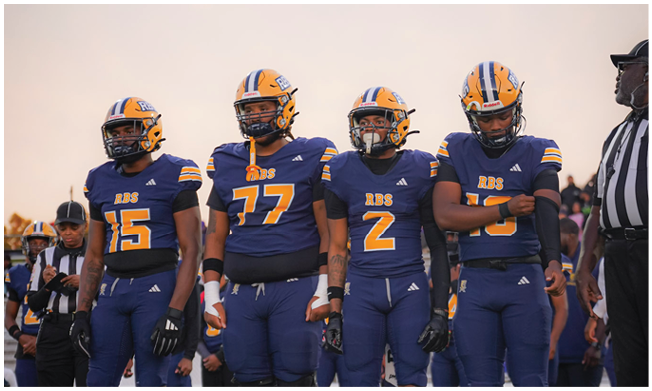 Four people wearing yellow and blue football helmets stand outdoors under a clear sky with a fifth person wearing a cap and glasses standing to the right. Description generated by AI