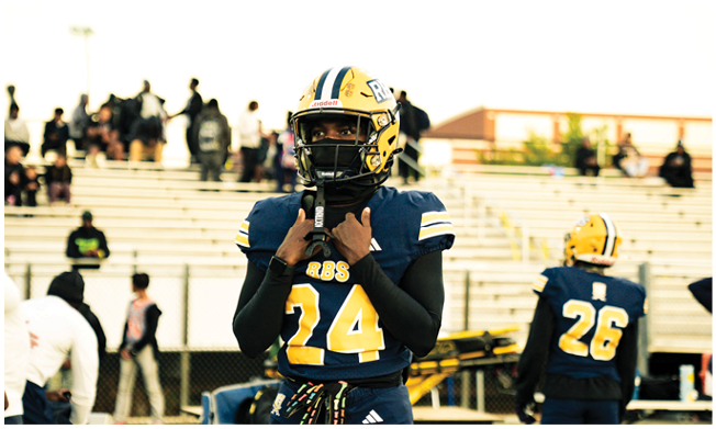 A person wearing a blue and yellow football uniform with the number 24 stands on a field adjusting their helmet while another person in a similar uniform with the number 26 stands nearby with bleachers and spectators in the background. Description generated by AI