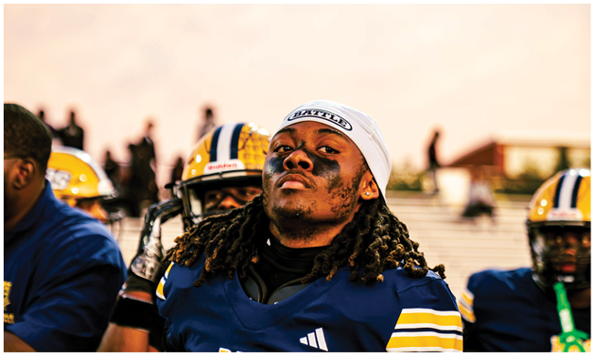 A person wearing a football uniform and a white headband looks confidently at the camera while sitting on a bench with teammates in the background. Description generated by AI