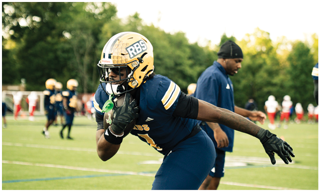 A person in a blue football uniform and gold helmet is running on a field while holding a football, with other players and a coach in the background. Description generated by AI