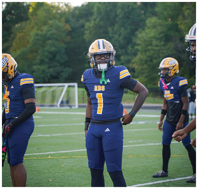 Three people wearing blue and yellow football uniforms and helmets stand on a field with trees in the background. Description generated by AI