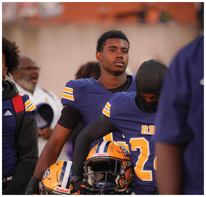 A person wearing a blue and yellow sports uniform stands among others in similar attire on a field with a blurred background. Description generated by AI