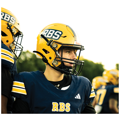 Two people wearing yellow helmets and navy blue football uniforms with “RBS" on the front are standing outdoors, with one person looking to the side. Description generated by AI