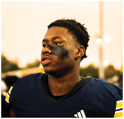 A person wearing a sports jersey with black face paint under their eyes looks to the side against a blurred outdoor background. Description generated by AI